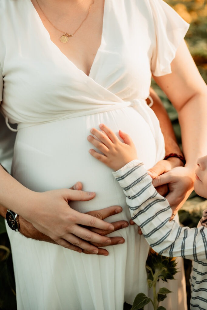 Séance maternité en extérieur en Alsace : le grand frère pose sa main sur le ventre de bébé, moment tendre en famille
