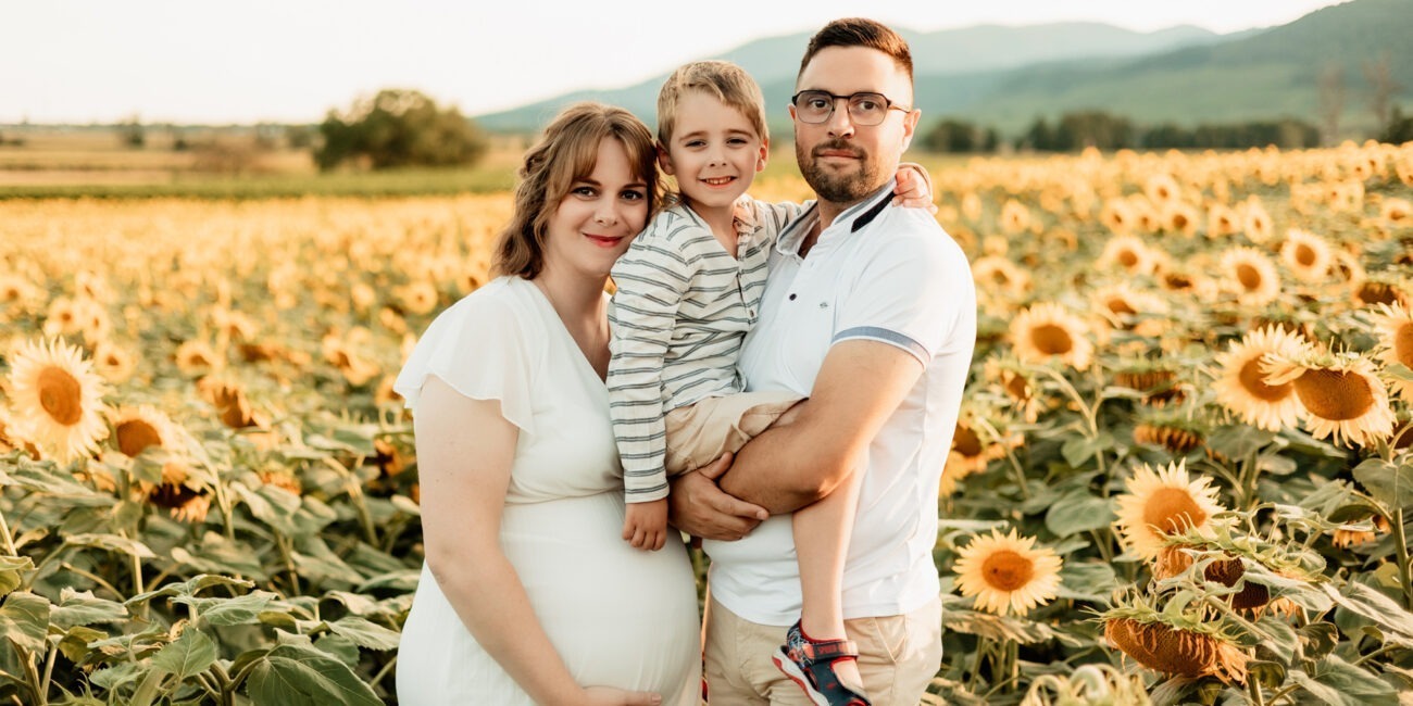 Séance maternité en famille en extérieur en Alsace : parents et grand frère dans un champ de tournesols au Bollenberg